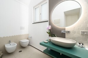 A modern bathroom with a round mirror, a bidet, a toilet, and a large vessel sink on a teal countertop. - Blue Sky Luxury Palace – Superior Orange Room | Elegant Ground-Floor Room (Meta)