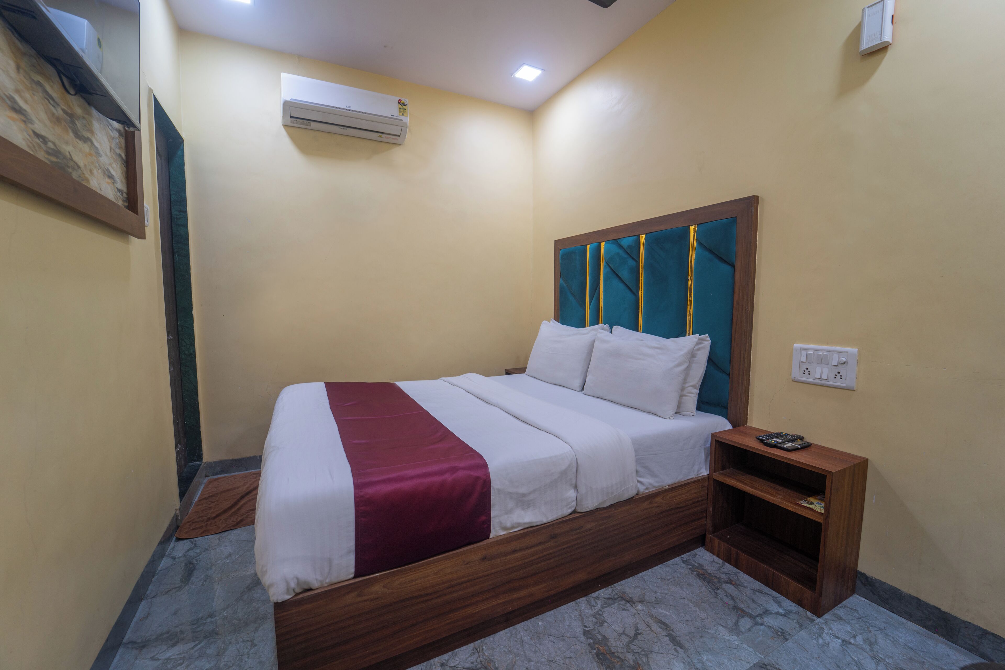 A bedroom with a wooden bed, white bedding, a red accent, a nightstand, and an air conditioning unit on the wall.