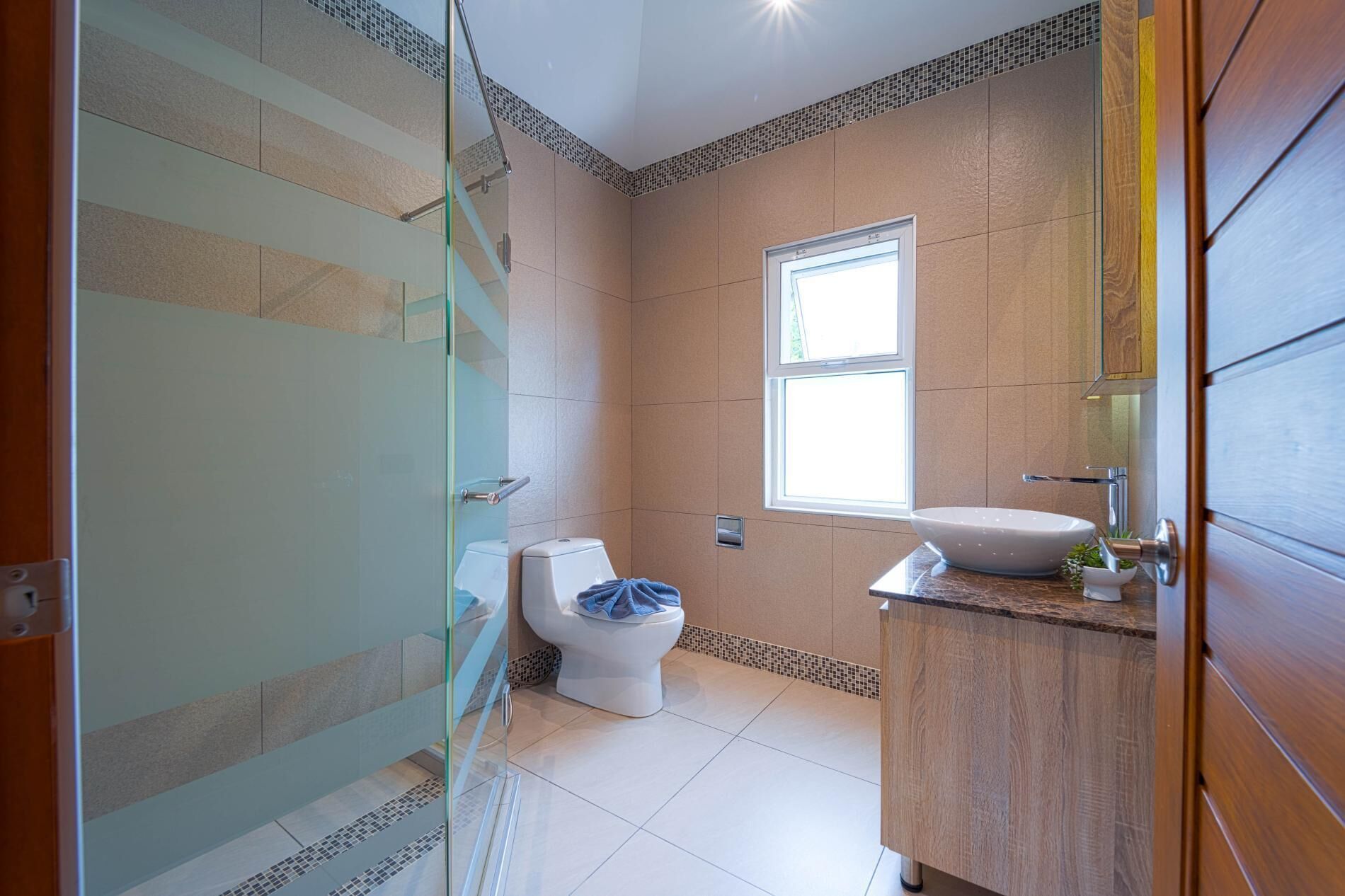 Une salle de bain moderne dotée d’une douche à parois de verre, de toilettes, d’un lavabo avec un miroir et d’une fenêtre laissant entrer la lumière naturelle.