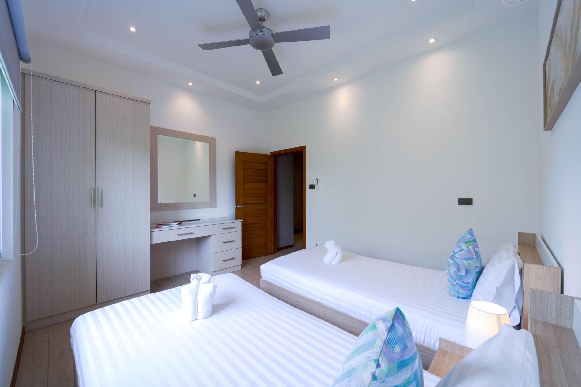 A hotel room with two beds, a wardrobe, a vanity with a mirror, a ceiling fan, and a door leading to another room.