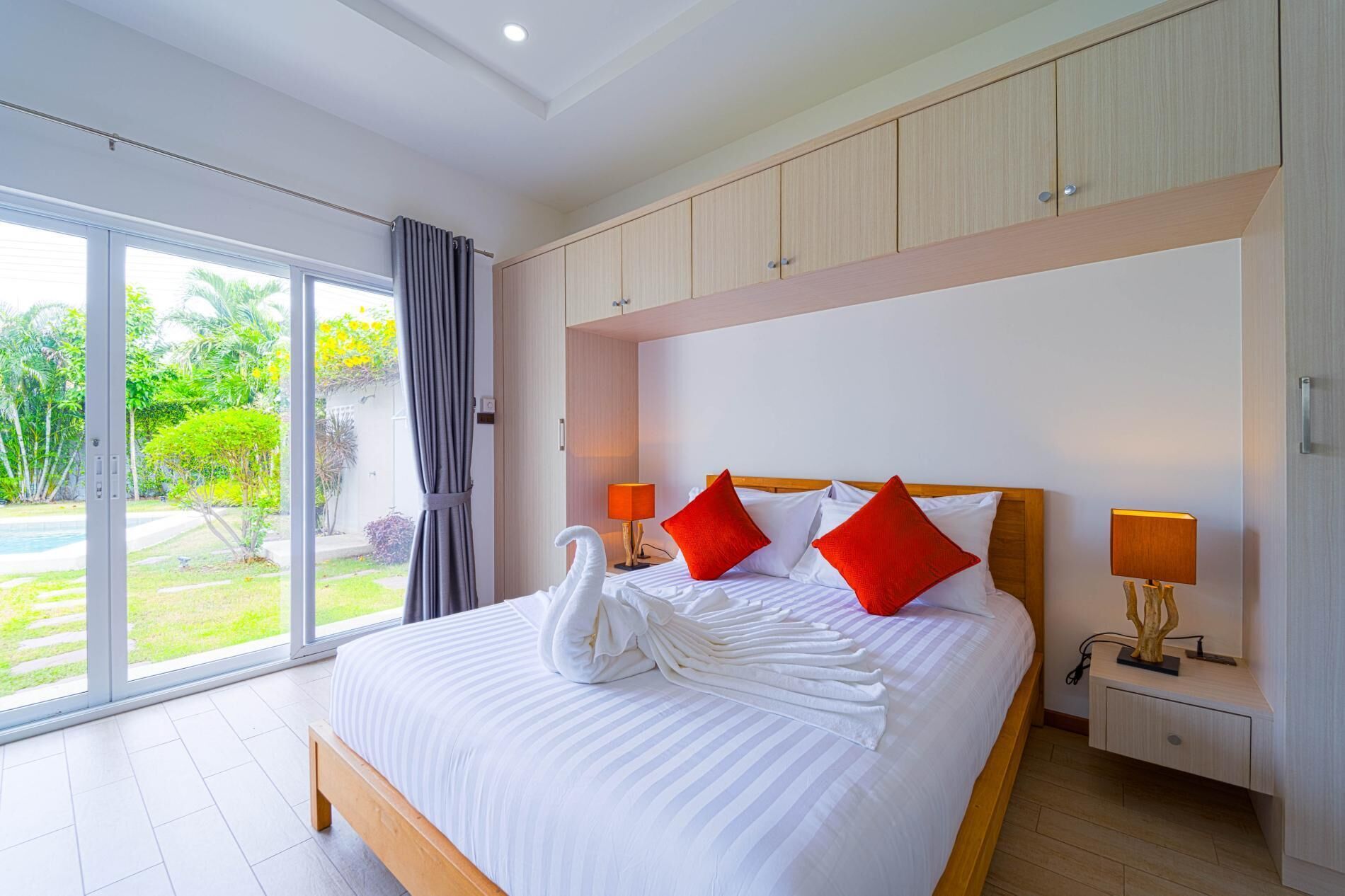 A hotel room with a large bed, two nightstands, a wooden headboard, and a view of a garden through a sliding glass door.