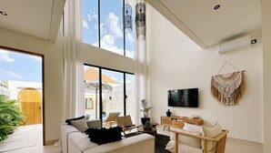 A modern living room with a large sofa, a coffee table, and a TV mounted on the wall. There is a large window providing a view of the outside. - Villa Sylvia Canggu (Canggu)