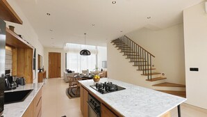 A modern kitchen with a central island, wooden staircase, and a dining area with natural light. - Villa Sylvia Canggu (Canggu)