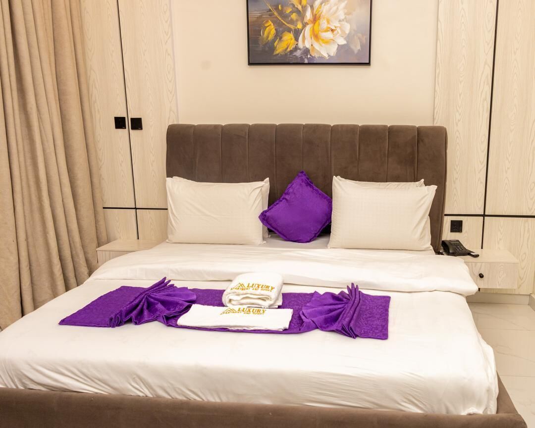 A neatly made bed with white linens and purple accents, a painting of flowers on the wall, and a wooden wardrobe with a light finish.