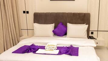 A neatly made bed with white linens and purple accents, a painting of flowers on the wall, and a wooden wardrobe with a light finish.