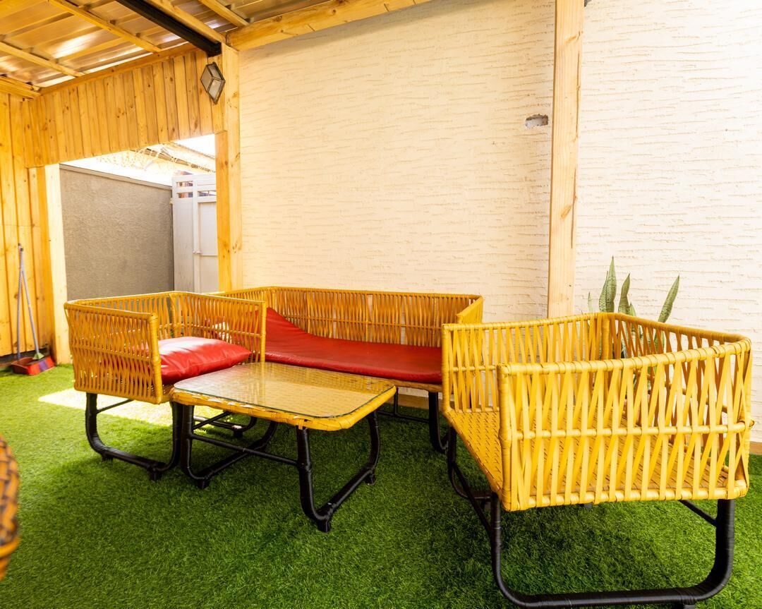Une pièce avec un sol recouvert d’un gazon artificiel vert, un canapé en osier avec des coussins rouges et une petite table.