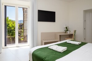 A hotel room with a bed, a TV mounted on the wall, a small desk with a chair, and a balcony with a view of greenery. - Blue Sky Luxury Palace | Apartment with Private Jacuzzi near the Beach (Meta)