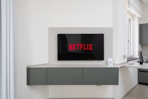 A modern living room with a large flat-screen TV mounted on a white wall, a grey cabinet below, and a window to the right. - Blue Sky Luxury Palace | Apartment with Private Jacuzzi near the Beach (Meta)