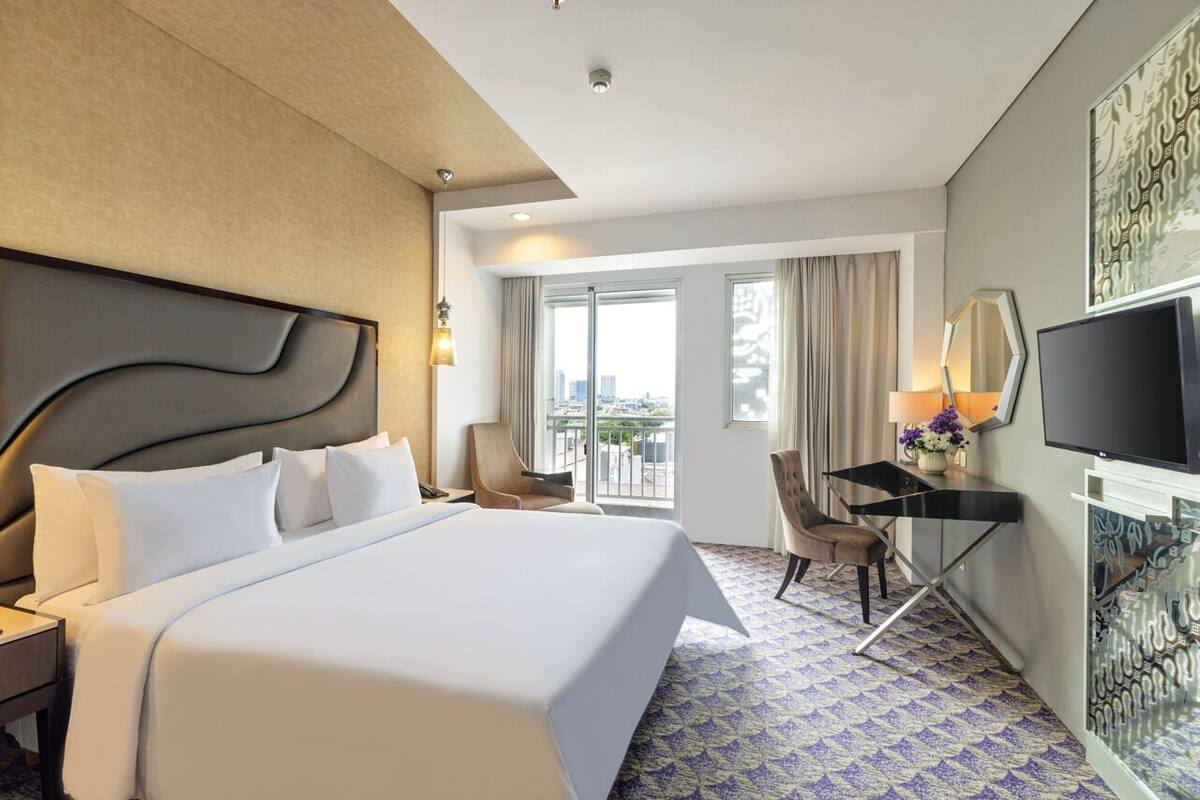 A hotel room with a large bed, a desk, a chair, a TV, and a balcony with a city view.