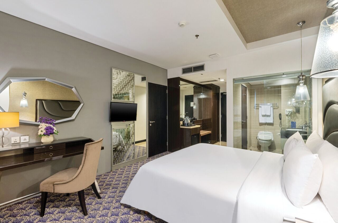 A hotel room with a bed, a desk, a chair, a mirror, and a bathroom visible through a glass partition.