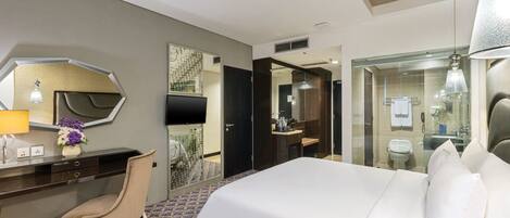 A hotel room with a bed, a desk, a chair, a mirror, and a bathroom visible through a glass partition.