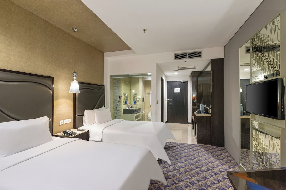 A hotel room with a large bed, bedside table, lamp, TV, and a bathroom visible through an open door.