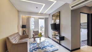 A modern hotel room with a sofa, a coffee table, a TV, and a large window with curtains. - Fairview Hotel by ARTOTEL (Jakarta)