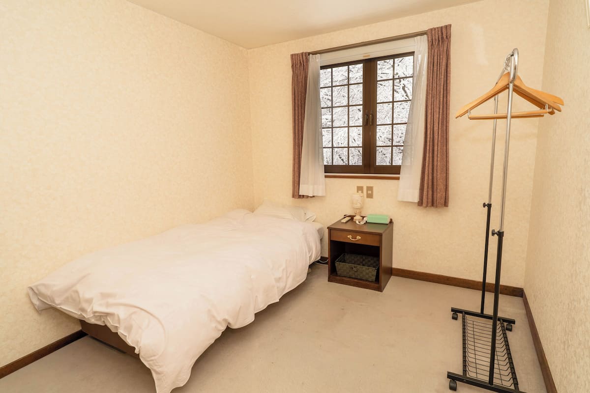 Een kleine, verzorgde slaapkamer met een enkel bed, een nachtkastje, een kledingrek en een raam met gordijnen.