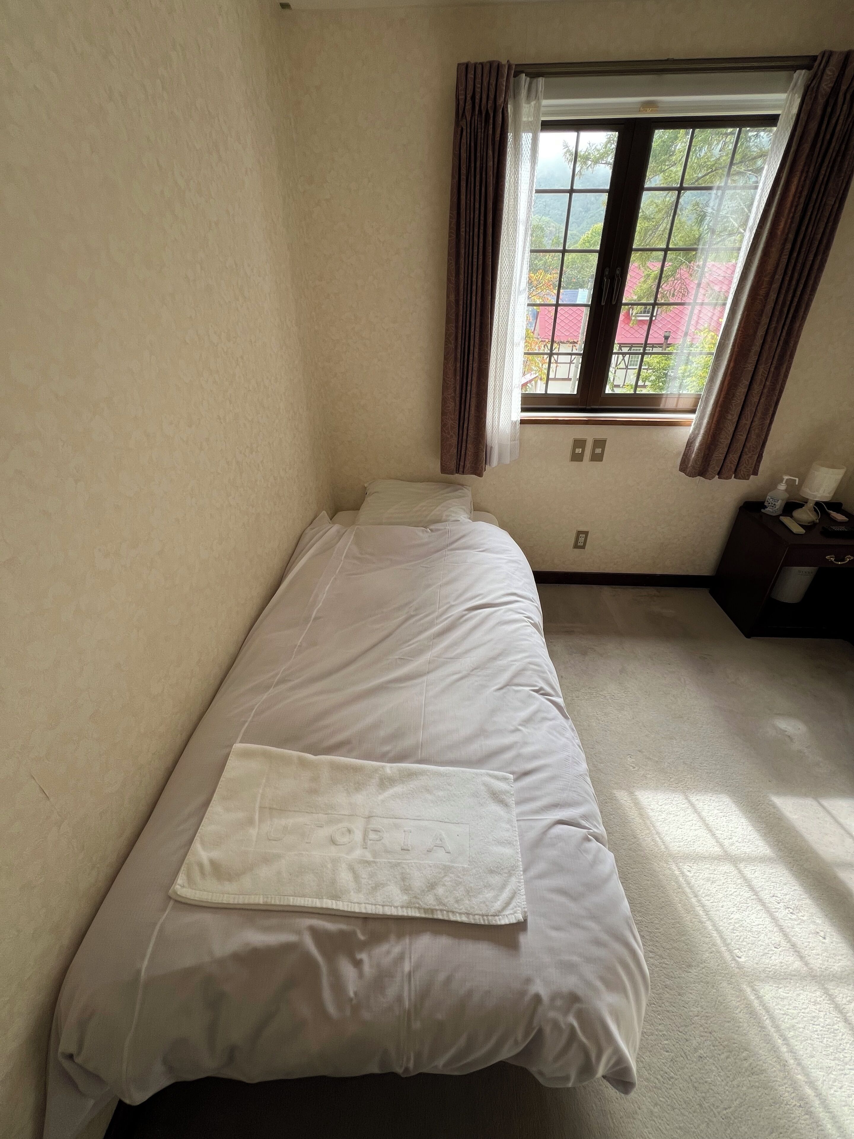 Ein Einzelbett mit weißer Matratze und Kissen, ein Fenster mit Vorhängen und ein kleiner Nachttisch mit Lampe und Flasche.