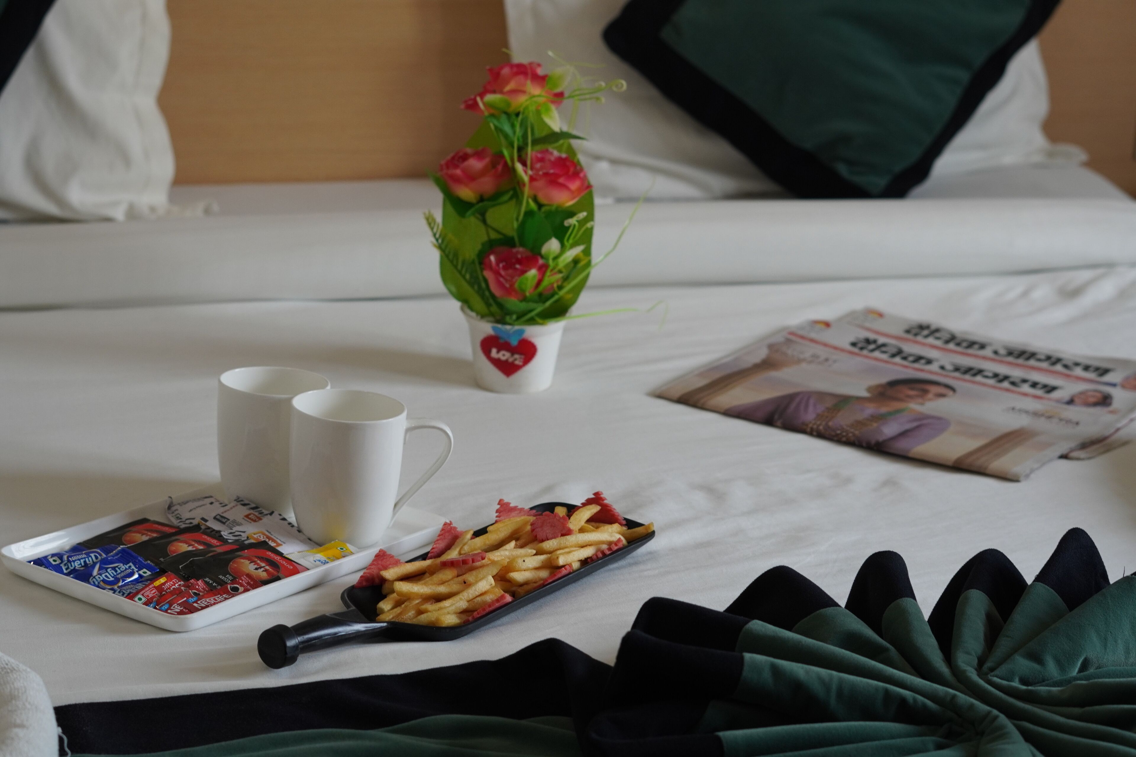 A neatly made bed with a magazine, a tray of food, a cup, and a vase of flowers.