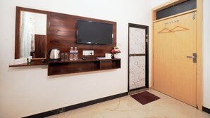 A reception area with a flat-screen TV mounted on a wooden panel, a coffee maker, water bottles, a small plant, and a wooden door with a coat hanger. - Hotel Kanak Kuteer (Ayodhya)