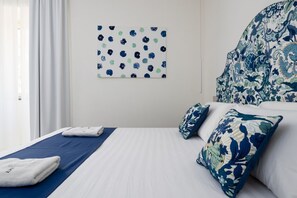 A hotel room with a bed, pillows, a blue and white patterned headboard, a wall art piece, and a window with curtains. - Blu Sky Luxury Palace – King Blue Apartment: Regal Comfort & Contemporary Design in Meta 👑💙 (Meta)