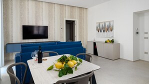 A modern dining area with a table set for two, a blue sofa, a flat-screen TV mounted on the wall, and a kitchen area with a white cabinet and a fruit display.