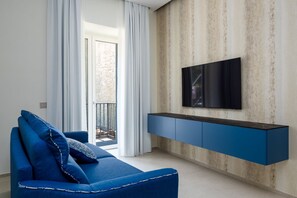 A modern living room with a blue sofa, a flat-screen TV mounted on a textured wall, and a balcony with white curtains. - Blu Sky Luxury Palace – King Blue Apartment: Regal Comfort & Contemporary Design in Meta 👑💙 (Meta)