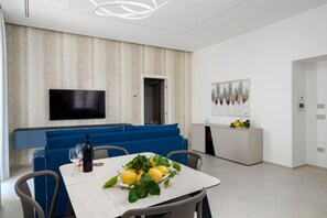 A modern dining area with a table set for two, a blue sofa, a flat-screen TV mounted on the wall, and a kitchen area with a white cabinet and a fruit display.
