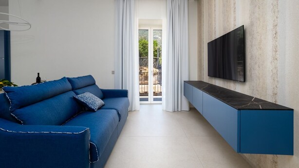 A modern living room with a blue sofa, a TV mounted on a textured wall, and a sliding glass door leading to an outdoor area.