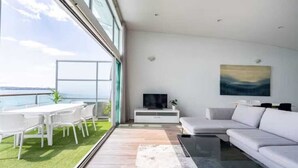 A modern living room with a large window, a white sofa, a coffee table, and a flat-screen TV. - Spacious Penthouse With Spectacular Harbour Views! (Auckland)