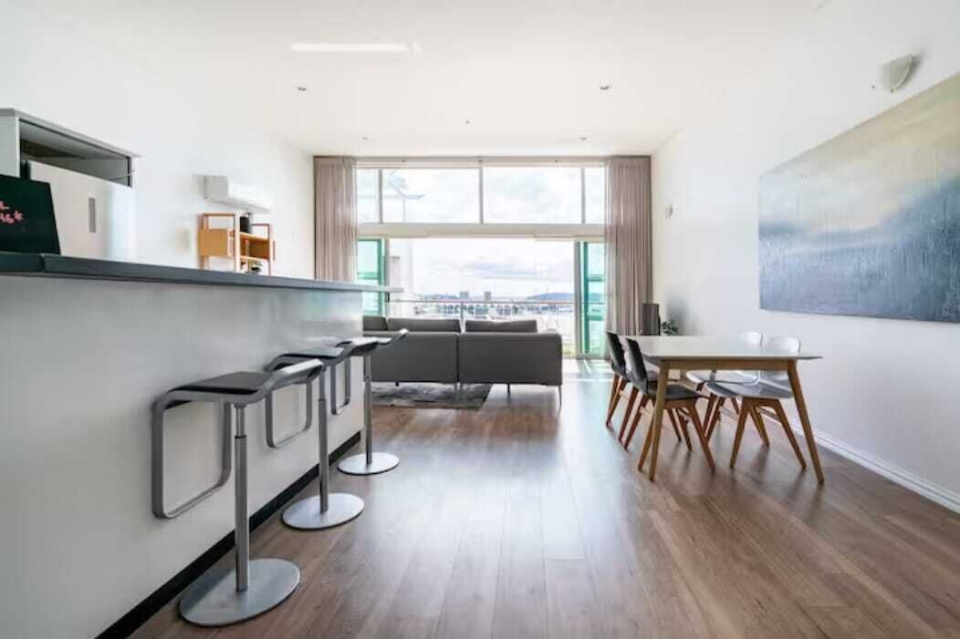 A modern kitchen with a dining area, a sofa, and a large window with a city view.