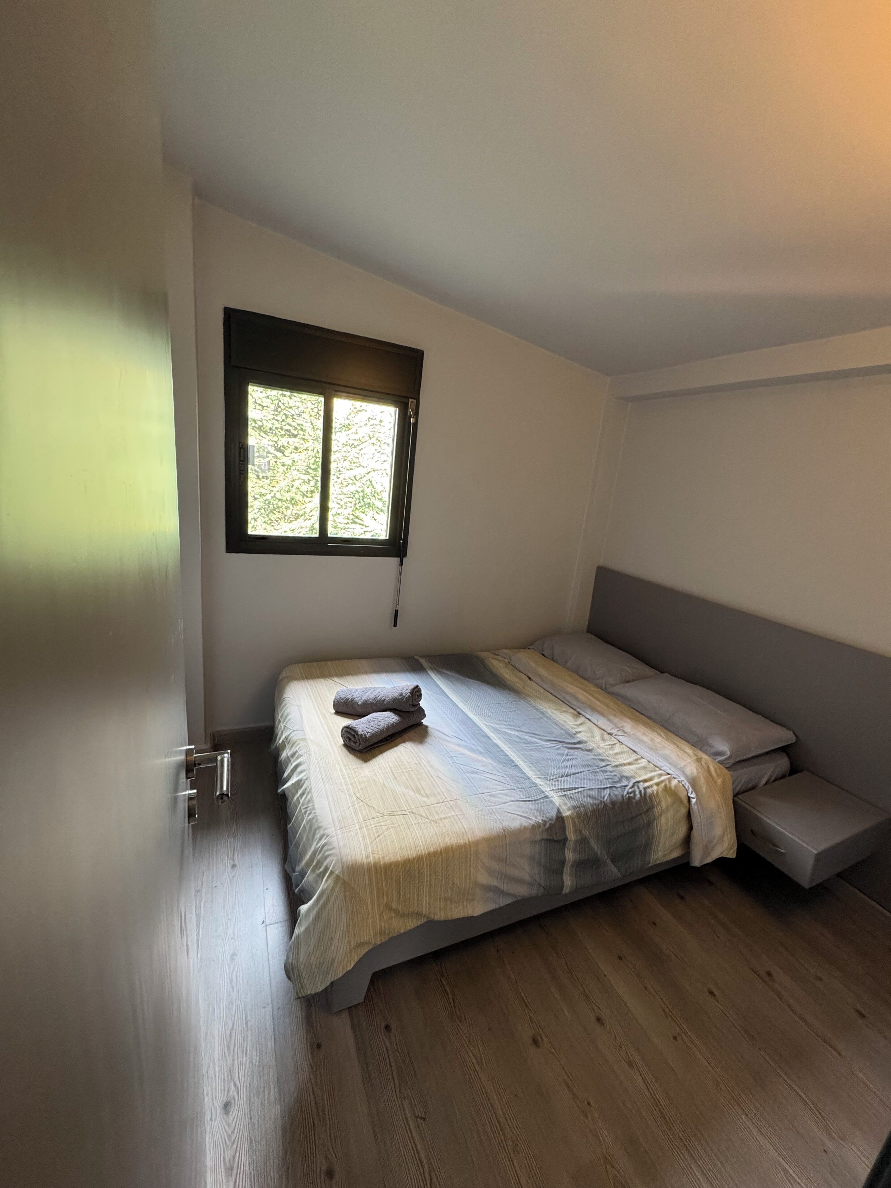 A small, modern bedroom with a single bed, a window, and a wooden floor.