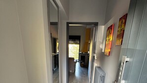 A hallway with a wooden floor, a grey door, and a radiator. There is a mat at the entrance and a painting on the wall. - The Cozy Inn - Agate (Faqra - Kfardebiane,)
