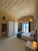 Un dormitorio con paredes de piedra, una cama con colcha azul, una mesita de noche con lámpara y vistas al mar a través de una ventana.