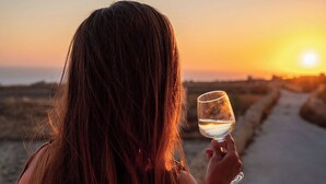 Una persona sosteniendo una copa de vino blanco en una terraza de madera con vista a una playa al atardecer.