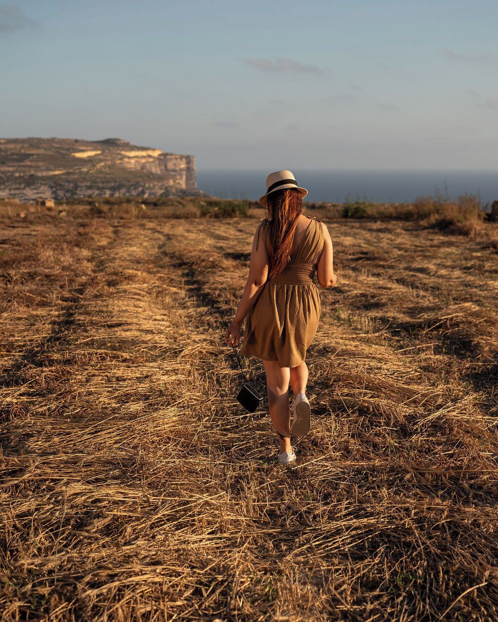 Una persona caminando sobre un campo seco y cubierto de hierba, con un acantilado al fondo.