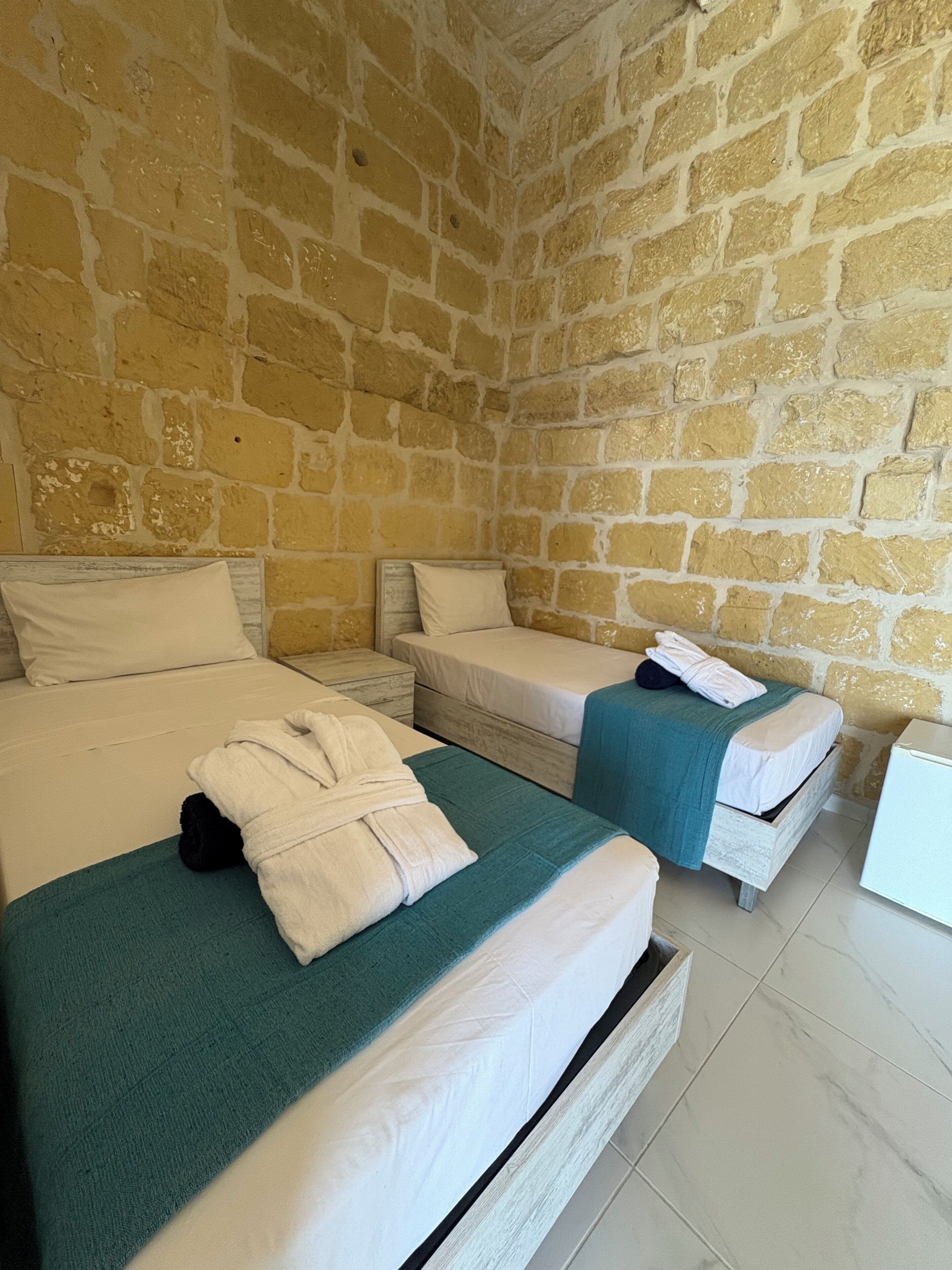 A room with two beds, each with a white mattress and teal bedspread. The walls are made of stone, and there is a white nightstand between the beds.