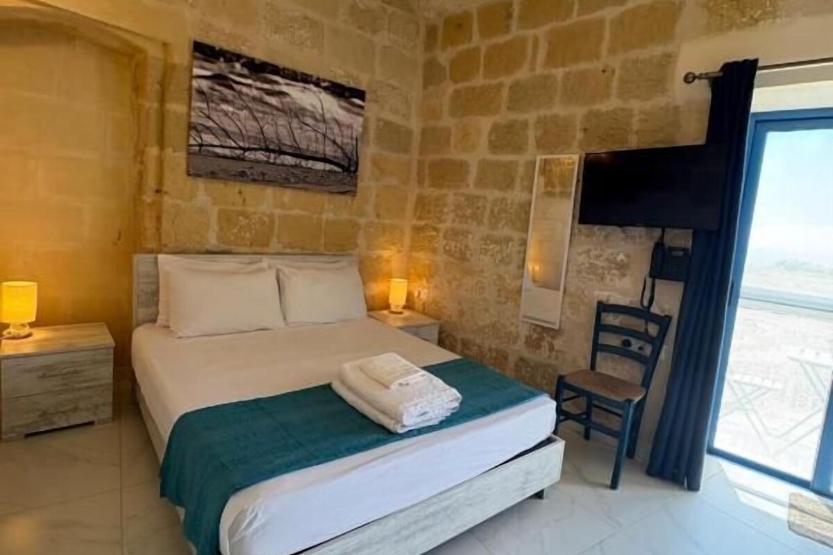 A bedroom with a stone wall, a bed with white linens, a nightstand, a chair, and a window with curtains.