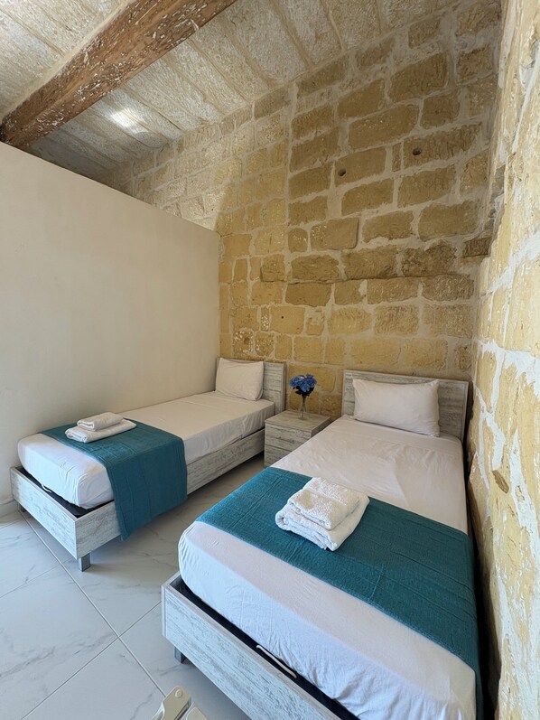 A bedroom with two single beds, each with a white mattress and teal blanket. The room has stone walls and a wooden ceiling.