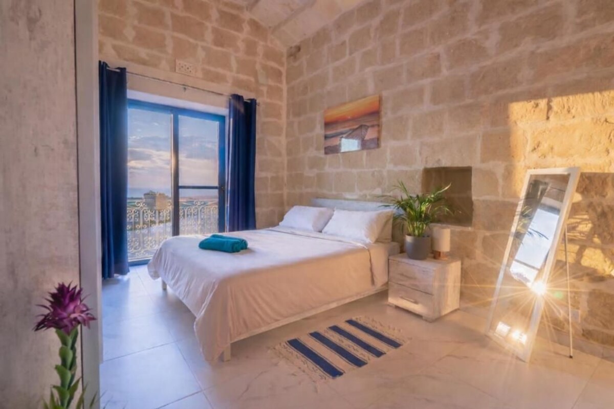 A bedroom with a bed, a nightstand, a potted plant, a framed picture, and a view through a window.