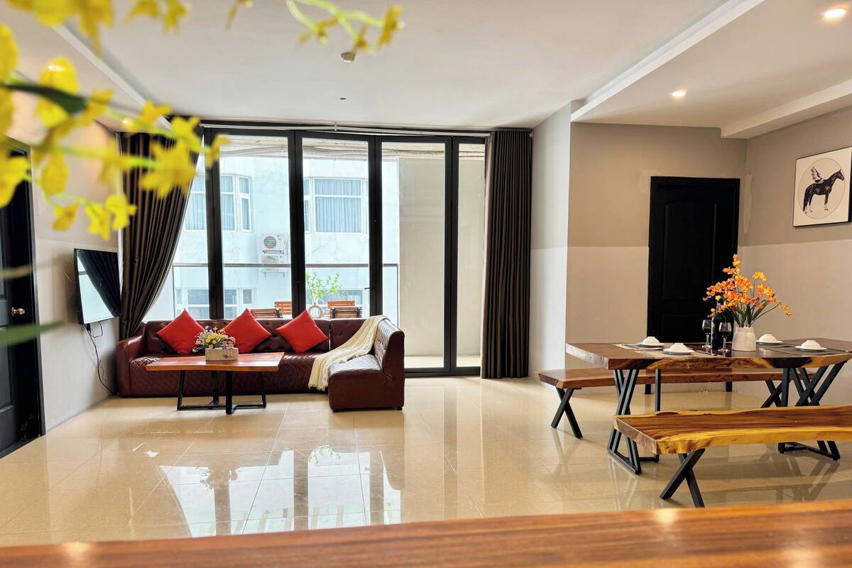 A modern living room with a leather sofa, a wooden dining table, and a large window with curtains.
