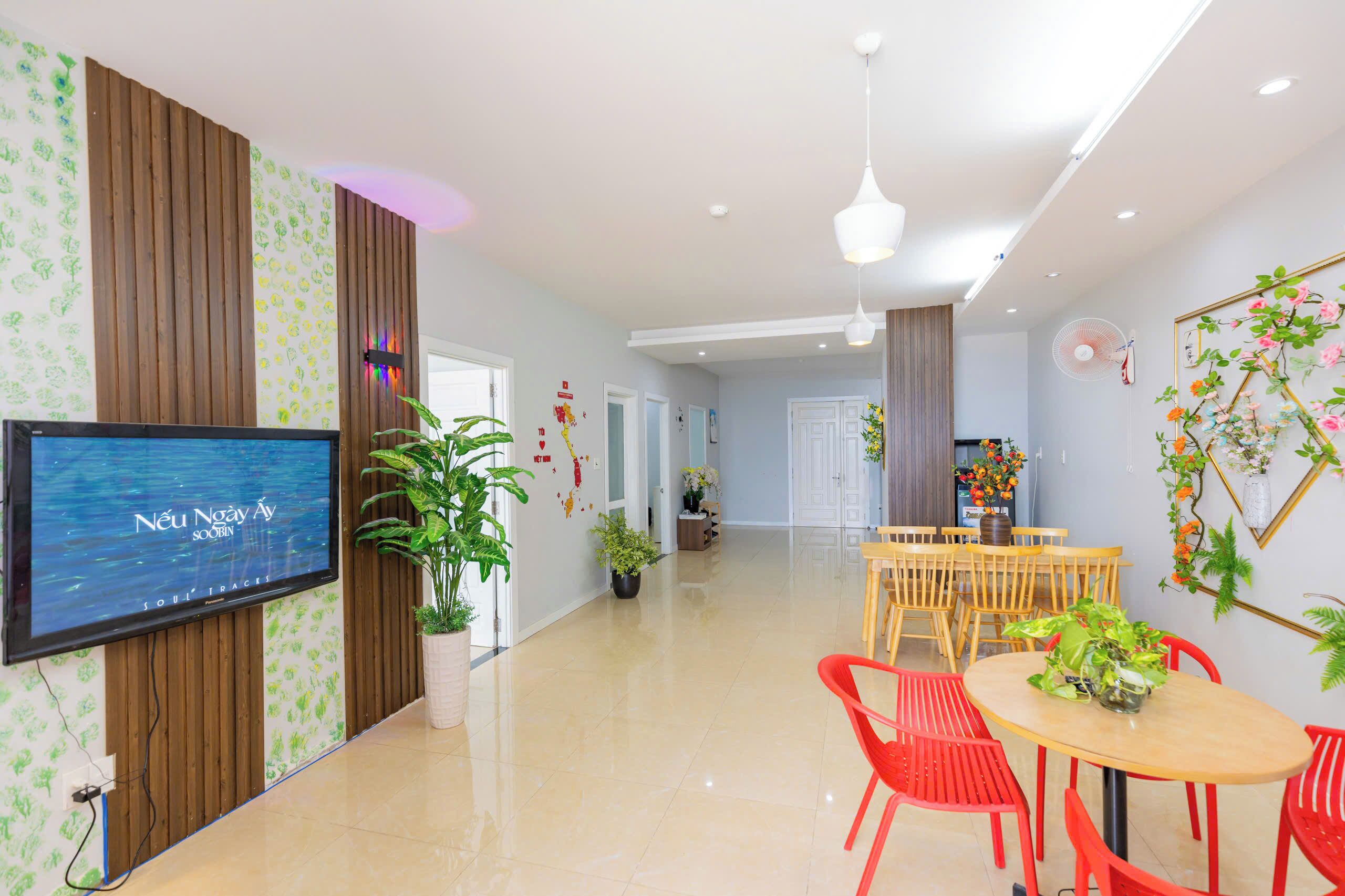 Un interior moderno con televisión de pantalla plana, paneles de madera, una zona de comedor con sillas rojas y plantas en macetas.