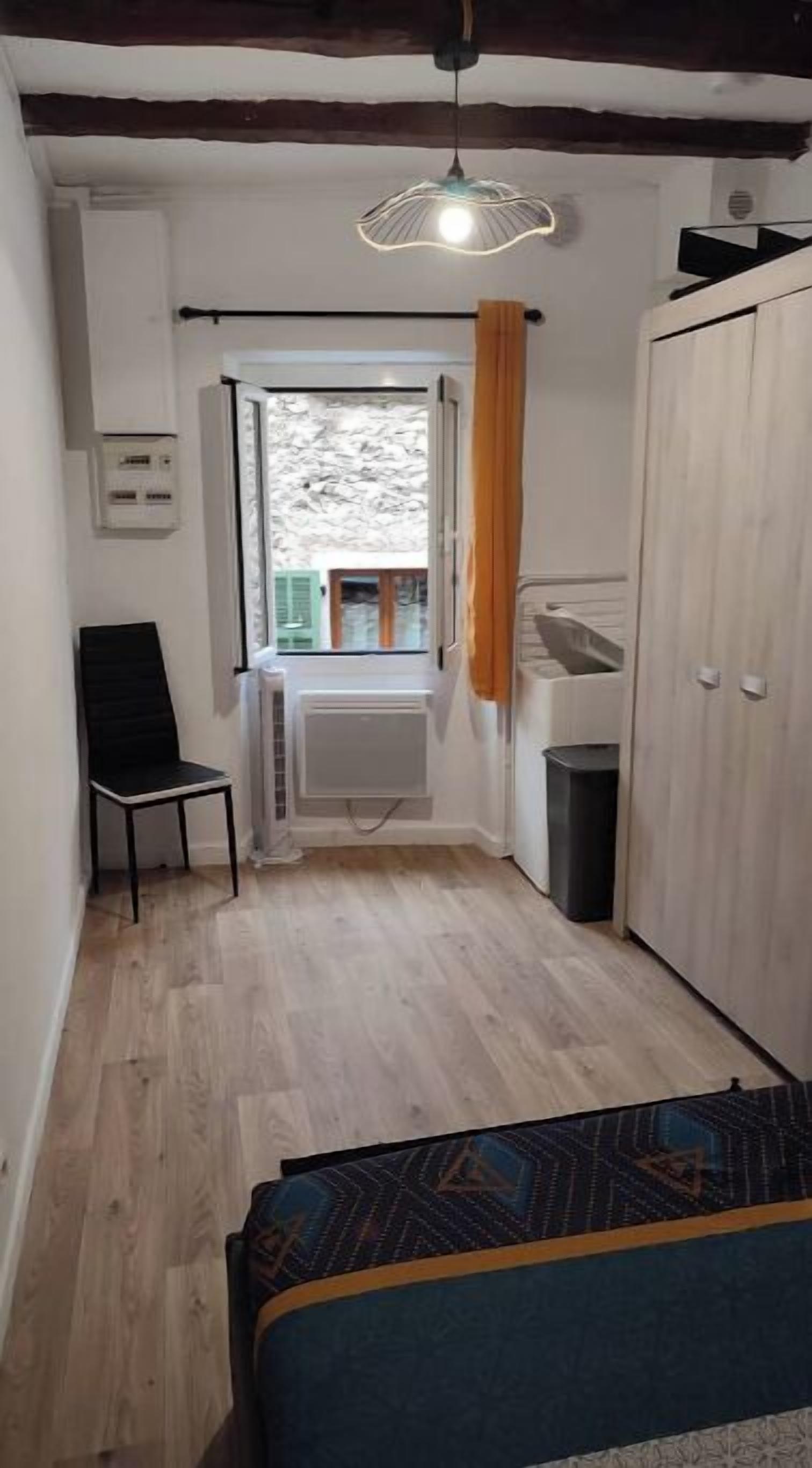 Une petite pièce étroite, avec un lit, une chaise, un radiateur, une fenêtre avec des rideaux et une armoire.