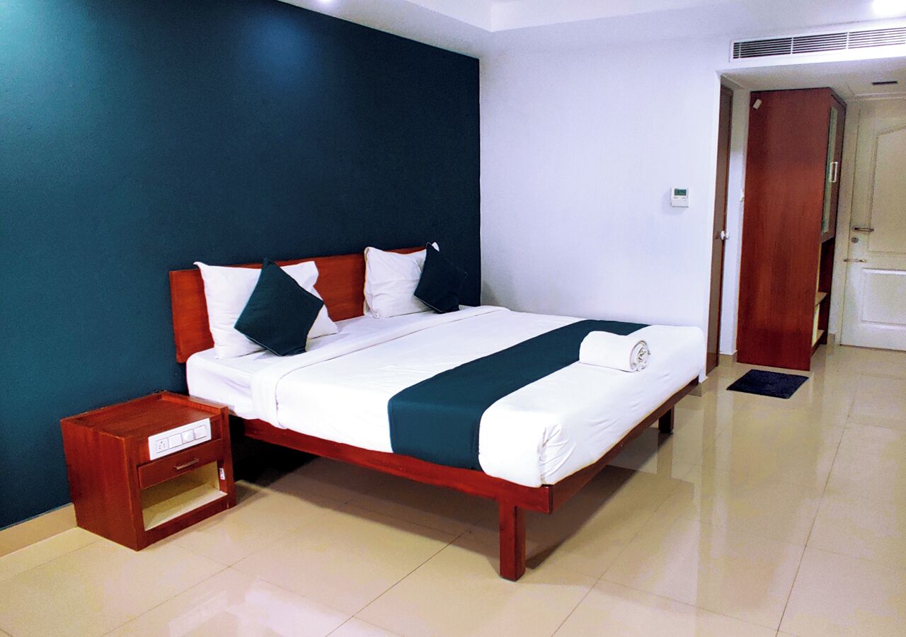 A neatly made bed with a wooden frame and white bedding, a small wooden nightstand with a drawer, and a door leading to another room.