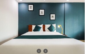 A neatly made bed with a white and teal bedspread, a wooden headboard, and teal and white pillows. There are framed pictures on the wall above the bed. - The Reach Hotel (Ernakulam)