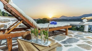 A wooden lounge chair with a cushion, a tray with two glasses of a clear beverage with mint, and a blue towel on a stone-tiled floor with a sunset view. - Villa Simantiri - Romantic Luxury Villa with jacuzzi & sunset views (Pompia)