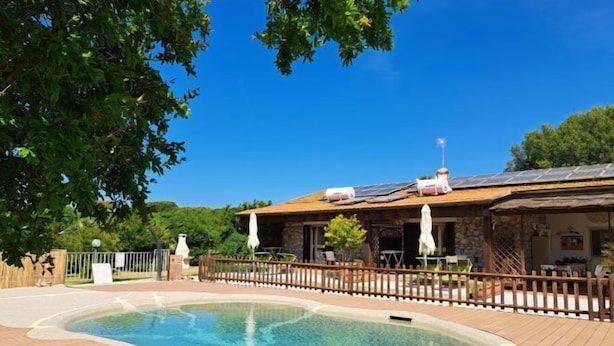 Une piscine avec une terrasse en bois, un bâtiment en pierre doté d’un toit en tuiles et des panneaux solaires.