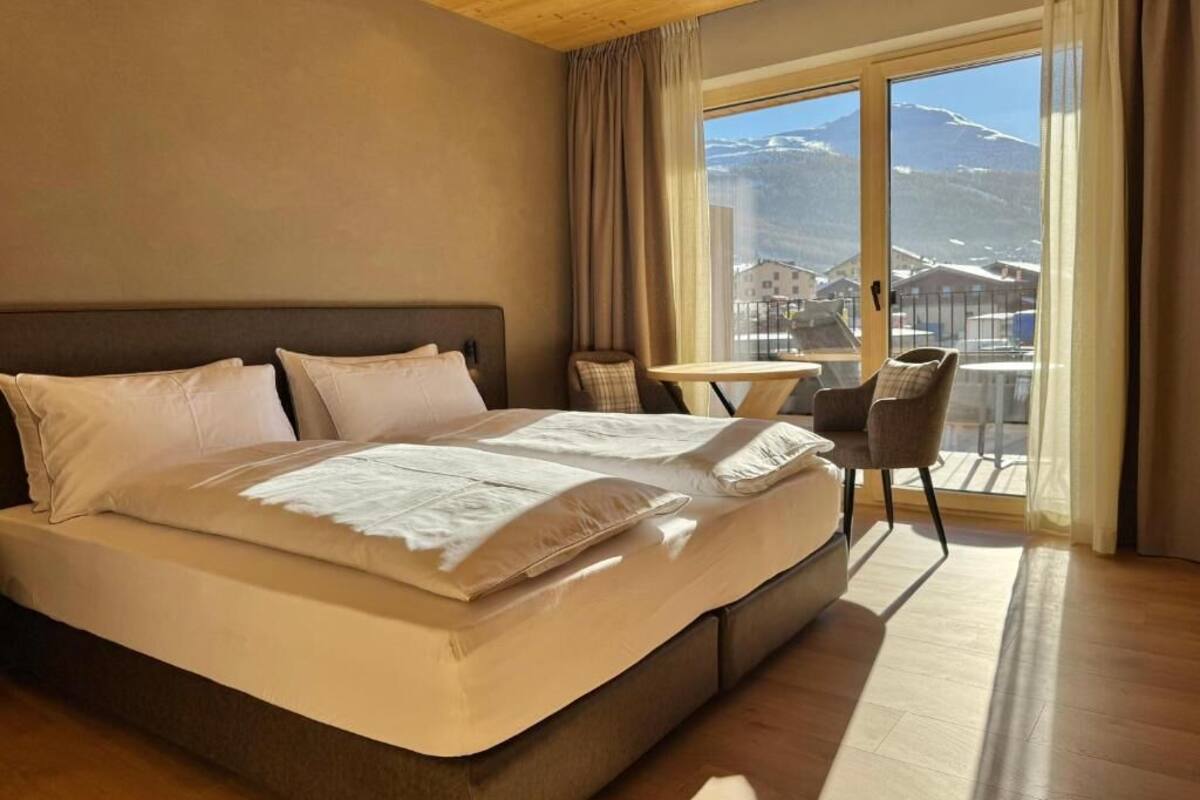 Un dormitorio con una cama grande, una mesita pequeña y una silla, y vistas a las montañas a través de un ventanal.