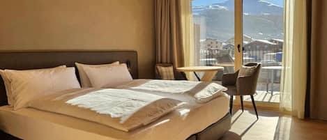 Un dormitorio con una cama grande, una mesita pequeña y una silla, y vistas a las montañas a través de un ventanal.