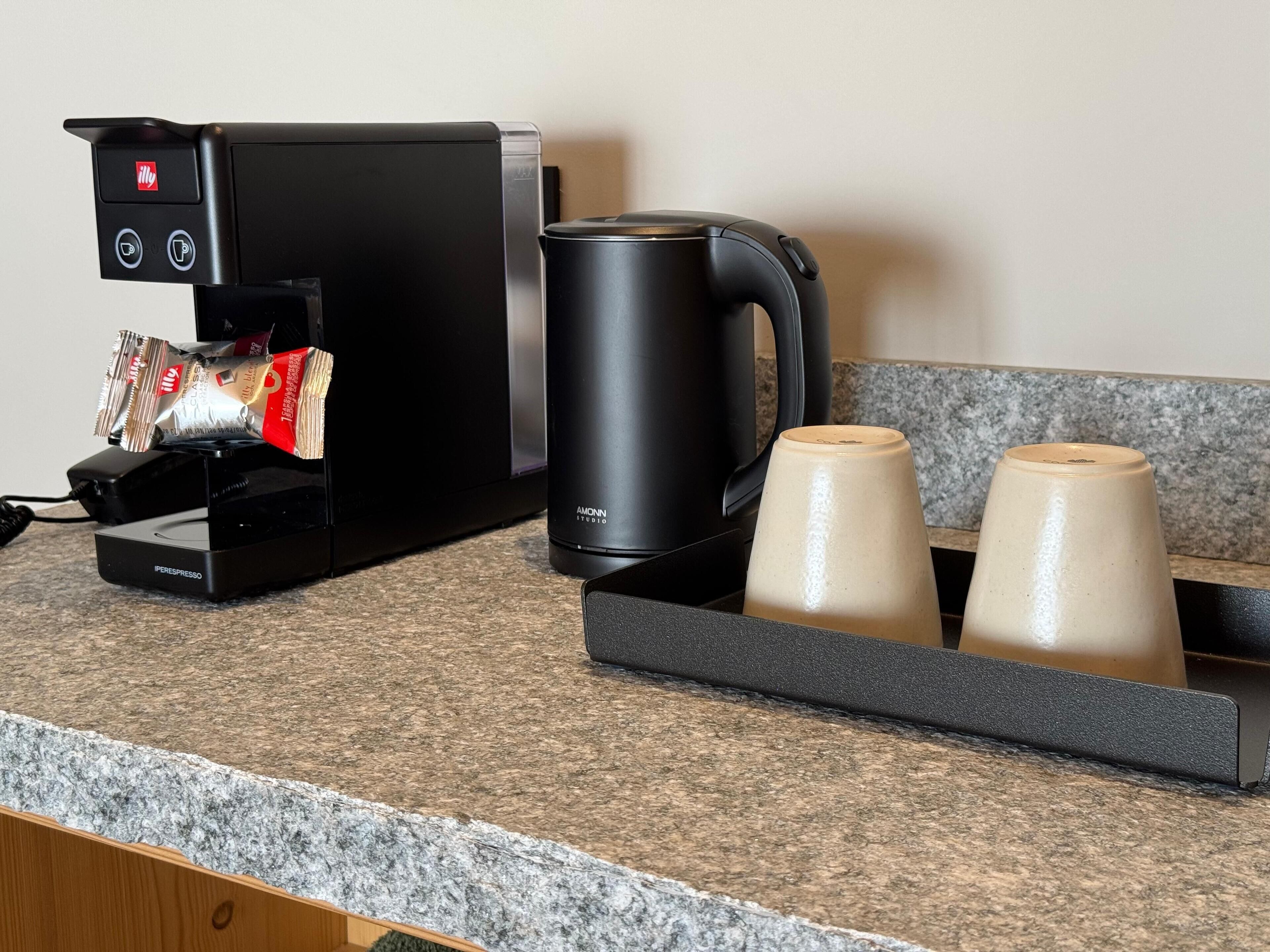 Una cafetera con dispensador de cápsulas, una máquina de café negra y dos tazas de cerámica sobre una encimera de granito.