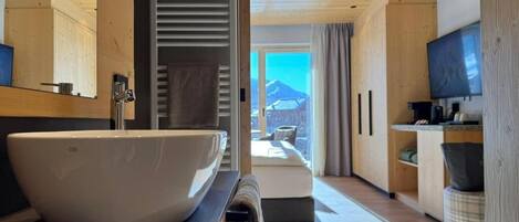 A modern bathroom with a large white basin, wooden flooring, and a view of a mountain through a large window.