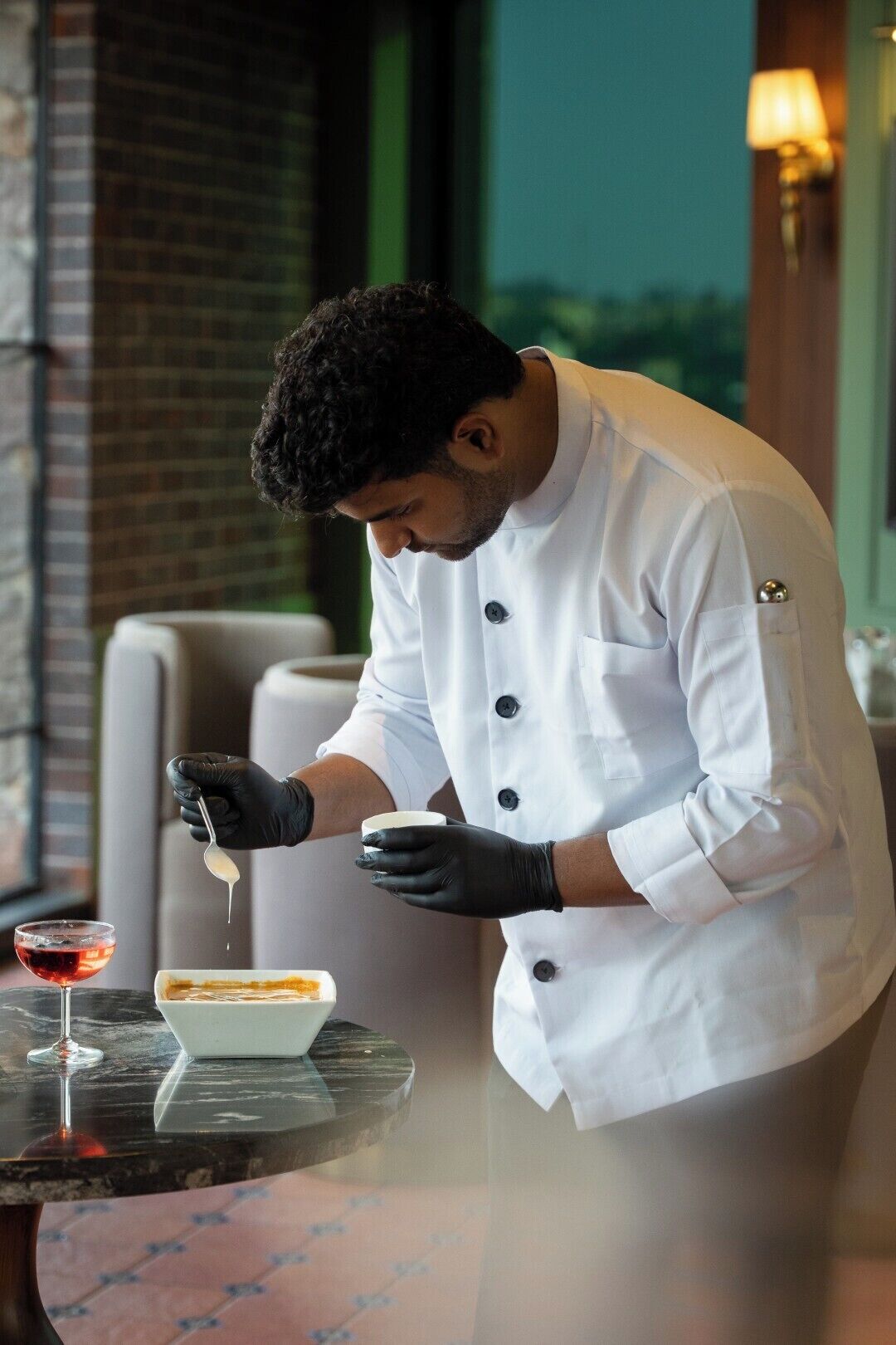 A chef in a white uniform and black gloves preparing a dish on a table with a glass of red wine and a dessert.