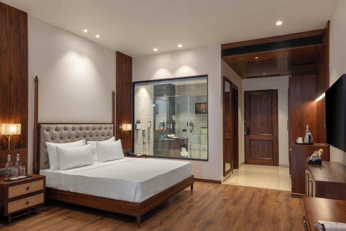 A modern hotel room with a large bed, wooden paneling, a glass shower enclosure, and a TV on a wooden cabinet.